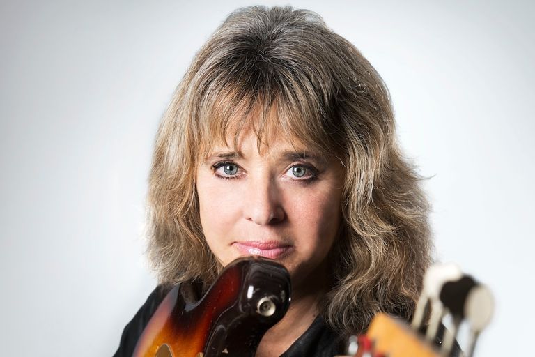 Rock icon Suzi Quatro posing for a close-up studio portrait while holding her electric bass guitar against a light background.